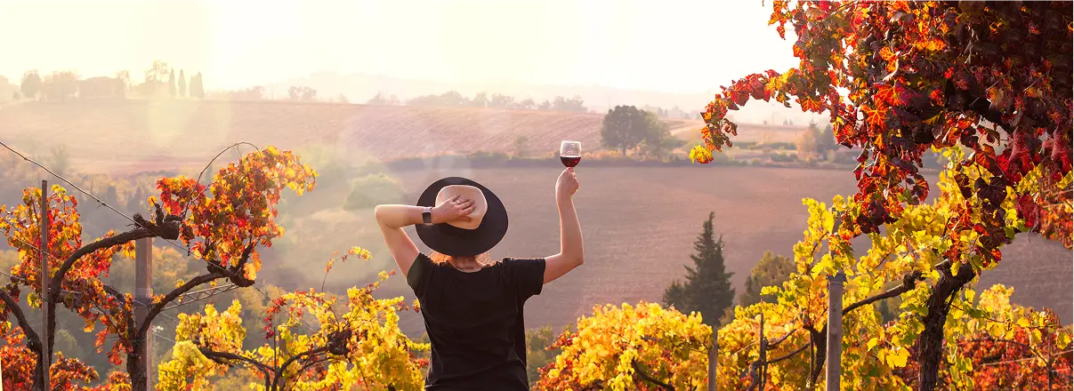 Donna con vino in vigneto toscano al tramonto. Colori autunnali, relax e destinazione di viaggio.
