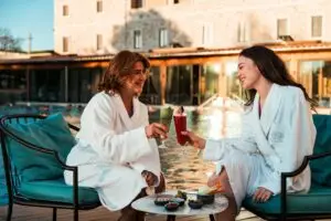 Due donne in accappatoio brindano a bordo piscina con cocktail colorati. Relax e benessere.