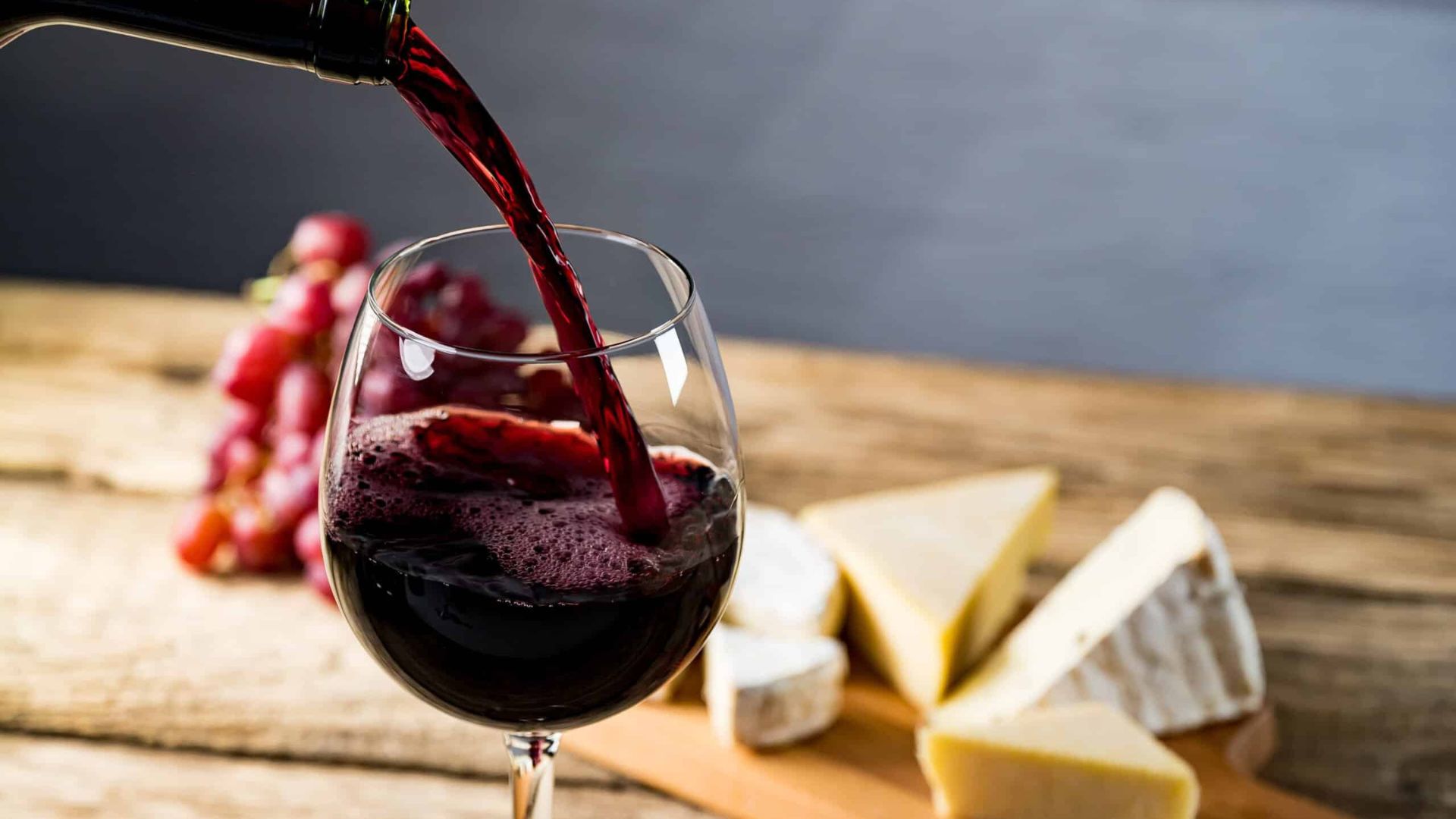 Versamento di vino rosso in un bicchiere con uva e formaggio sullo sfondo.